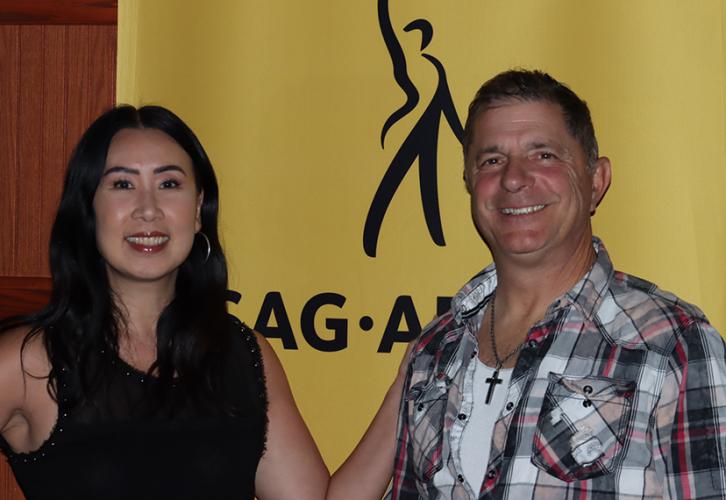 Kim, in black, and a male guest, wearing a plaid shirt, stand in front of a yellow SAG-AFTRA banner. 