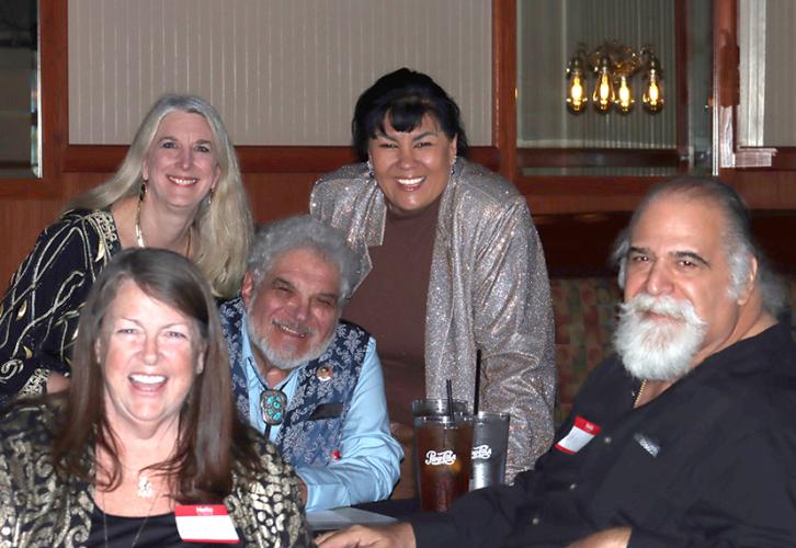 Two men and a woman sit at a table while two women stand behind them. There are drinks on the table.