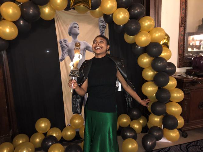 Williams, an African American woman, stands under a balloon arch. She wears a leather jacket, black shirt and green skirt. Her face is tilted back; her smile is wide. A photo booth prop, a gold paper statuette, is in her hand.