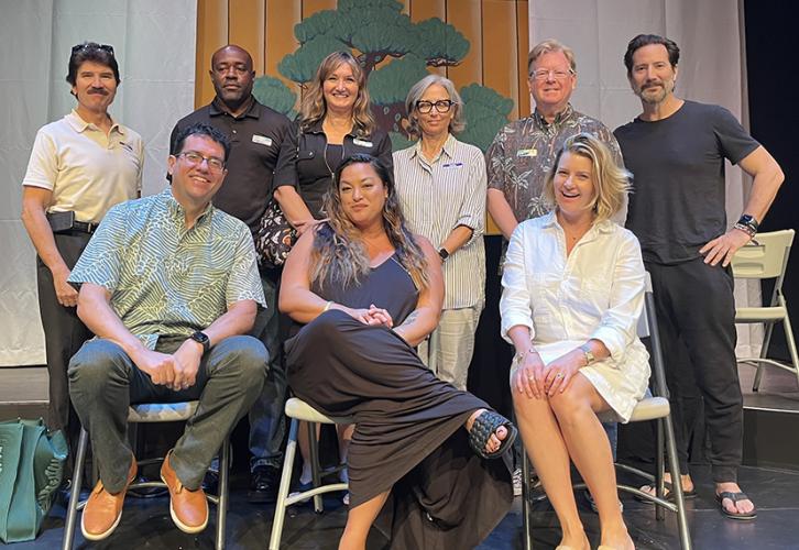 Nine people pose on a well lit stage in front of an orange backdrop with a bonsai tree.