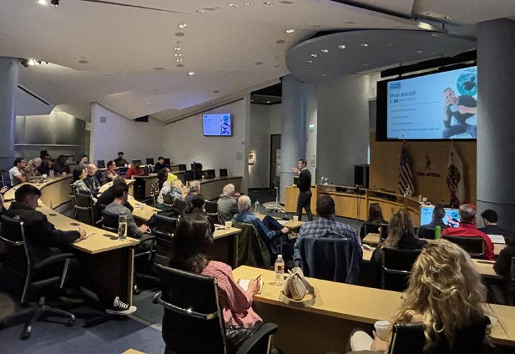 A room of individuals sit in lecture style seating while one individual teaches at the front.