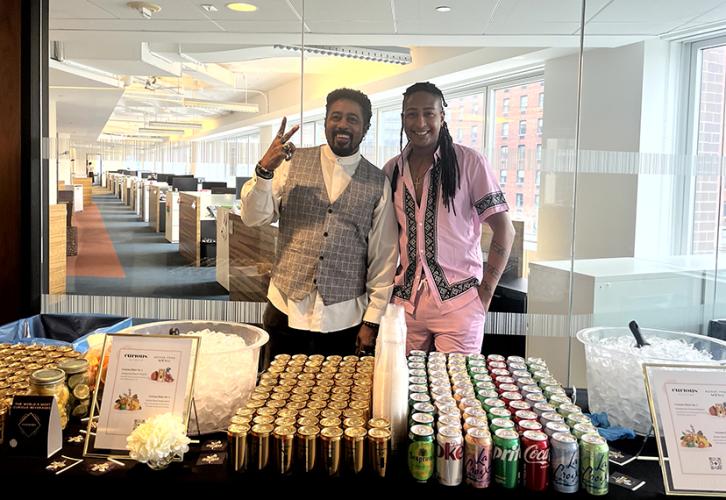 Two people stand behind cans of Curious Elixirs and soda.
