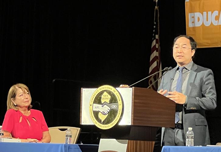 A woman is seated at a table to the left. A man stands at a podium with the New Jersey State AFL-CIO logo on the front in yellow and gray.