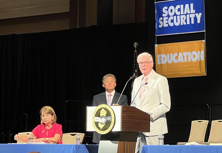 A woman is seated at a table to the left and a man stands in the background behind a podium. A man stands speaking at the podium with the New Jersey State AFL-CIO logo on the front in yellow and gray. Two banners hang behind him reading from the top.