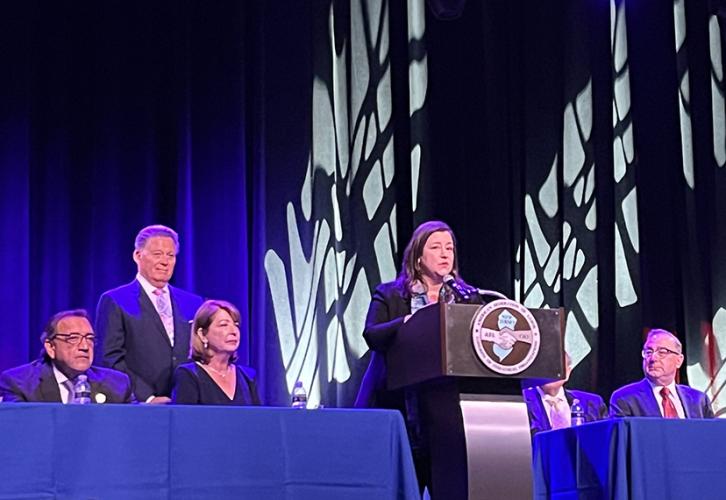 A woman stands at a podium addressing attendees. Four people are seated at a dais behind her with blue tablecloths. Lights reflect on the blue curtains behind them.