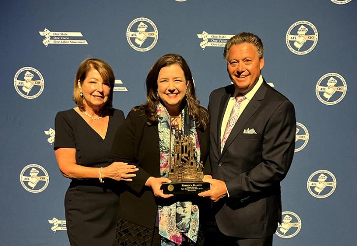 Two women and a man stand in front of a blue backdrop with the New Jersey State AFL-CIO logo in white. The woman in the center holds a metal award. 