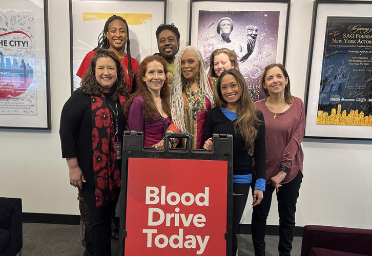 Seven women and one man stand in the SAG-AFTRA New York office behind a sign that reads "Blood Drive Today." There are four posters for SAG-AFTRA and the SAG-AFTRA Foundation on the wall behind them. 