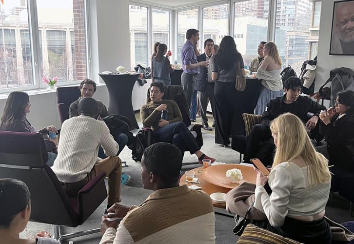 A diverse group of people are sitting in purple chairs and standing around black cocktail tables.