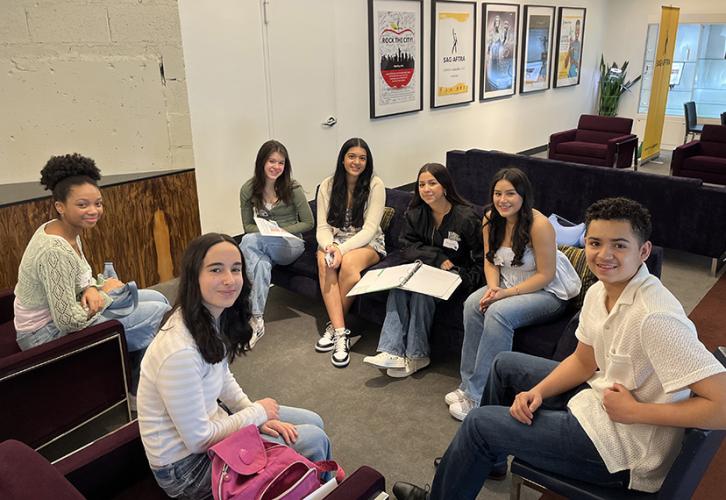 A diverse group of people are seated in a circle and smile for the camera. 