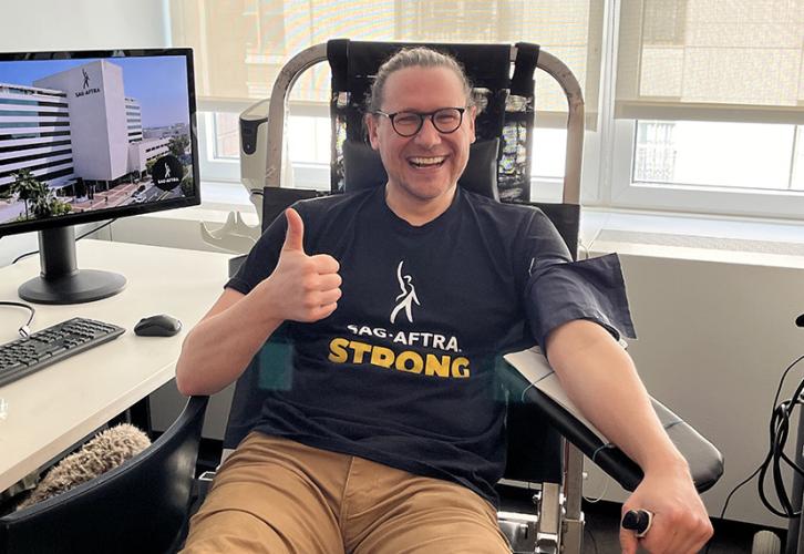 Thompson sits in a chair at a desk wearing a blood pressure sleeve. He is wearing a SAG-AFTRA Strong T-shirt and gives a thumbs-up sign.