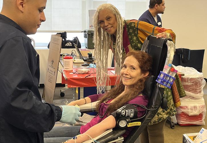 Lilly sits in a chair while a technician takes her blood. Black rests her hand on the back of the chair and leans into the picture.