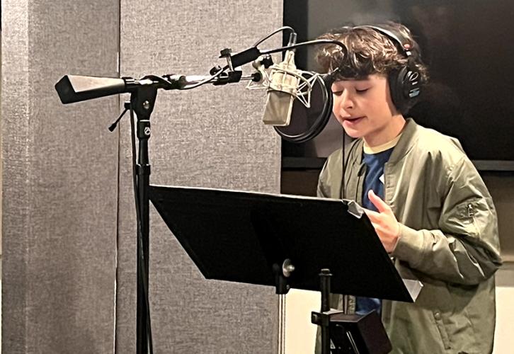 A child in a green jacket and black jeans stands behind a microphone and black stand. 