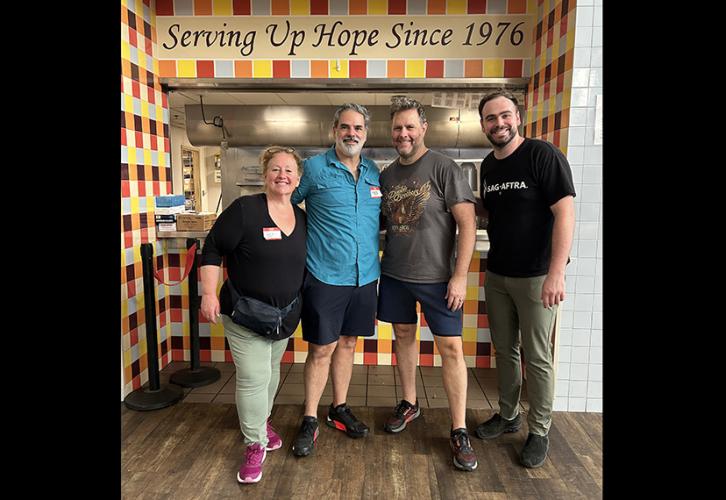 A woman and three men stand in an alcove adorned with colorful tiles. Above the doorway, it reads "Serving Up Hope Since 1976." All are dressed casually.