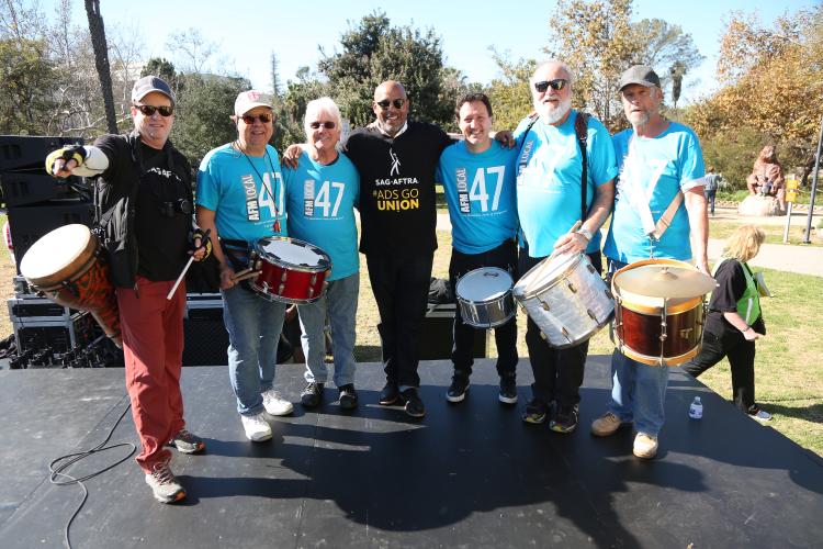 Group shot of 7 men on stage with their drums. 2 wearing SAG-AFTRA shirts and 5 wearing AFM Local 47 shirts.