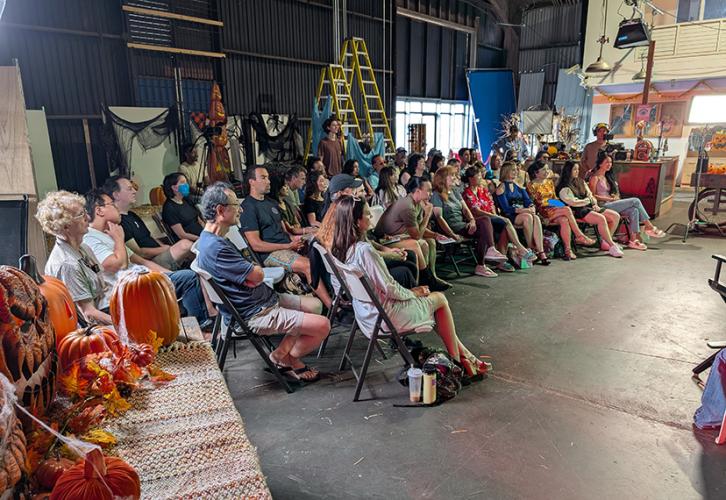 A diverse group of people sit in folding chairs in a semicircle on a film set. Lots of Halloween props adorn the space.