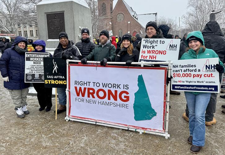 A group of four SAG-AFTRA New England Local staff members join three local union members and one IATSE member to rally against passing “right-to-work” legislation in New Hampshire. Four people hold individual signs and the group holds up a banner reading "Right to Work Is Wrong for New Hampshire.” All wear hats advertising SAG-AFTRA or IATSE and winter jackets.