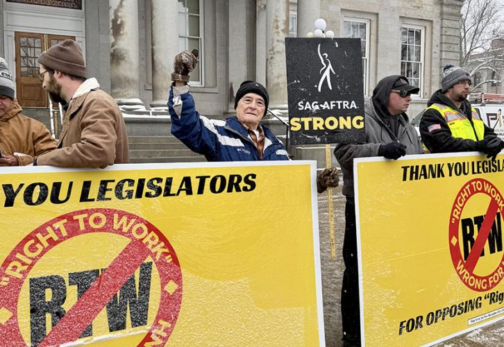 A SAG-AFTRA New England Local member raises a fist to protest right-to-work legislation in New Hampshire while holding a “SAG-AFTRA Strong” sign. Two bright yellow anti-right-to-work banners stand in front of him, held up by fellow protesters at the rally on Feb 13.