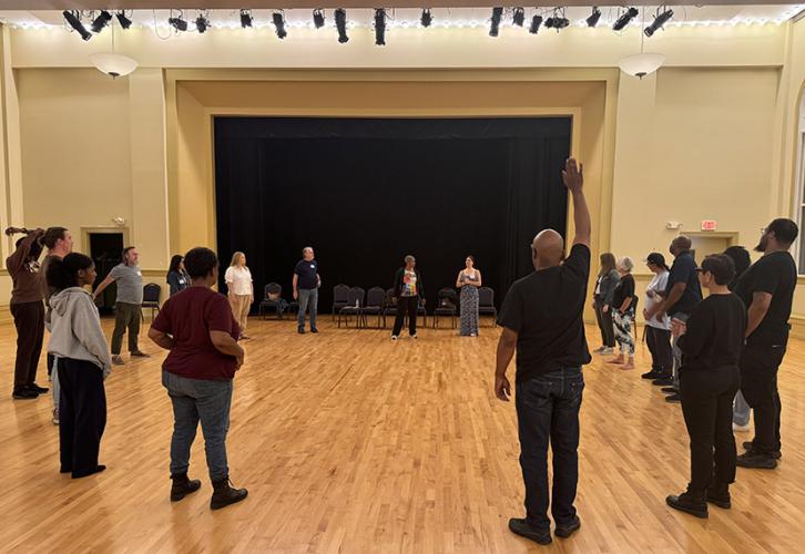 A diverse group of 20 people stand in a circle. One person has his right hand raised.