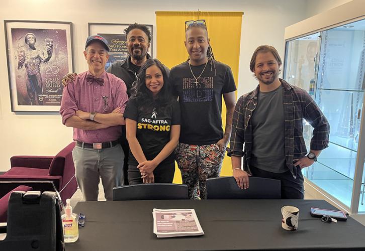 A diverse group of people stand behind a table with a black table cloth and two navy blue chairs. There is a stack of fliers on the table in front of them and two burgundy chairs on the left. There is a yellow SAG-AFTRA banner behind them.