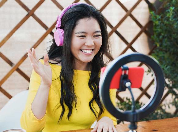 Image of female in yellow top wearing headphones looking at mobile with ring light behind.