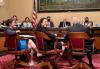 Long (Asian American) sits in a chair while smiling over her shoulder. In the background, senators sit behind a dais.