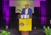 Knight (Black) is captured mid-speech on stage behind a podium bearing the Coalition of Black Trade Unionists’ name and logo. He wears a blue blazer and a light-colored dress shirt. He is framed by two fern plants and the lighting behind him is purple. 