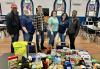 SAG-AFTRA New England Local Board member Gilda James, far right, and husband, New England Local member Chris James, far left, with staff of the Veterans Northeast Outreach Center in Haverhill after delivering donated items offered by members. Six people, three men and three women, stand in a church behind a table piled with food. There are three stained glass windows in the background.