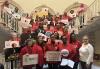 A diverse group of people wearing red and holding varying signs which read, “Sag-Aftra Strong,” “PowHer,” “Stronger Laws Now! Pay Transparency and Data Reporting,” “#EqualPayNY” are gathered around a staircase. 