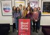 Seven women and one man stand in the SAG-AFTRA New York office behind a sign that reads "Blood Drive Today." There are four posters for SAG-AFTRA and the SAG-AFTRA Foundation on the wall behind them. 