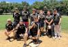 The SAG-AFTRA New York Local softball team after their win against Actors Federal Credit Union. A group of 13 softball players pose on a softball field. The players are wearing black jerseys with a yellow SAG-AFTRA logo in the center. There are green trees and a blue sky in the background.