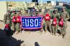 A woman in a blue sweatshirt and winter cap, center, stands with eight Army personnel in uniform. In the center is a blue USO flag with a red and white border and six white stars. They are outside. Hero image for "Giving Back to Those Who Serve"