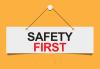 Safety First "Safety" in black "First" in red cursive on a white panel hanging from a whitepin with black string on a dandelion yellow background
