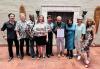 Nine diverse people pose in Honolulu Hale on the red brick floor, in front of a taupe trapezoidal wall with two commemorative koa wood surfboards, while the mayor holds the official framed proclamation.