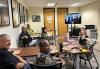 Performers learn about the SAG-AFTRA audiobooks contracts in the office boardroom and online via zoom. A group of diverse actors sit in a boardroom with a large screen filled with Zoom attendees at the front of the room; they smile at the camera. The carpet is standard office patterned gray, with off white walls adorned with framed movie posters. A large copier/printer/fax machine sits in the right side of the room with a trashcan near it. There are two wooden doors at the back of the boardroom.