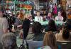 Five people sit at a table in folding chairs, smiling, on a set made up to look like a halloween store. In the foreground can be seen the backs of audience members.  