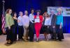 A diverse group of nine, seven women and two men, stand in a line on stage. Two women in the center hold a framed certificate. Behind them is a banner with illegible copy.