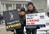 Two SAG-AFTRA New England Local members pose for a photo outside of the New Hampshire State Legislative Building. Both wear SAG-AFTRA hats and winter jackets and hold signs rallying against passing right-to-work legislation in the state.