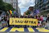 SAG-AFTRA New England Local members march across the historic Boston Marathon finish line on Boylston Street at the start of the June 8 Boston Pride for the People Parade. A large diverse group of people wearing and holding LGBTQIA+ SAG-AFTRA pride T-shirts and signs march on Boylston Street and cross over the finish line of the Boston Marathon as they participate in the Boston Pride Prade. People in the middle of the group hold a long, gold horizontal banner that reads “SAG-AFTRA.”