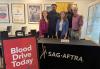 A diverse group of people stand behind a table covered in a black cloth with SAG-AFTRA logo  in white text. There is a red sign with Blood Drive Today in white next to the table.