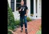 A man wearing blue jeans, boots and a black T-shirt holds a microphone while standing on a red brick sidewalk outside of a white wooden building.